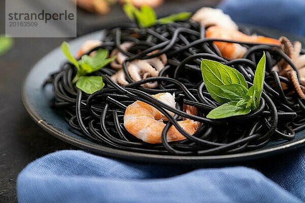 Schwarze Tintenfischnudeln mit Garnelen und kleinen Tintenfischen auf schwarzem Betonhintergrund und blauem Textil. Seitenansicht  Nahaufnahme  selektiver Fokus