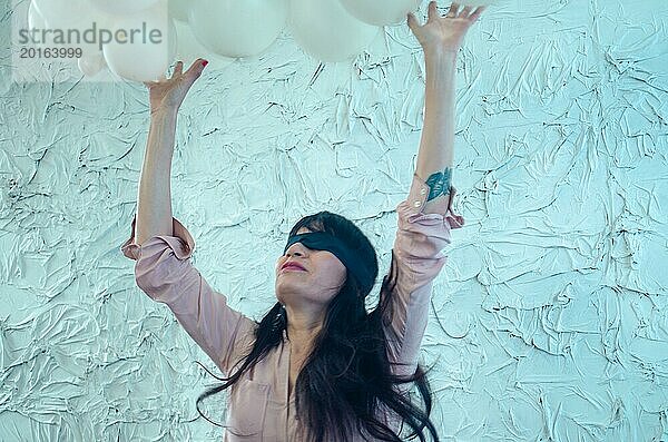 Eine spielerische junge erwachsene hispanische Frau mit verbundenen Augen  die sich in den Himmel streckt  umgeben von einer Fülle weißer Luftballons  künstlerische Performance  spielerischer Tanz