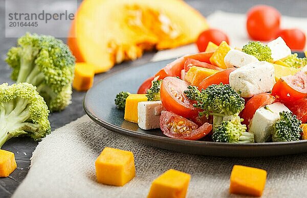 Vegetarischer Salat mit Brokkoli  Tomaten  Fetakäse und Kürbis auf einem blauen Keramikteller auf schwarzem Betonhintergrund und Leinentuch  Seitenansicht  Nahaufnahme  selektiver Fokus