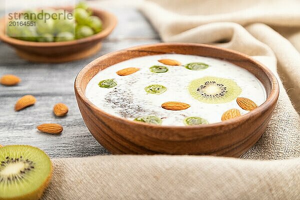 Joghurt mit Kiwi  Stachelbeere  Chia und Mandeln in einer Holzschale auf grauem Holzhintergrund und Leinenstoff. Seitenansicht  Nahaufnahme  selektiver Fokus