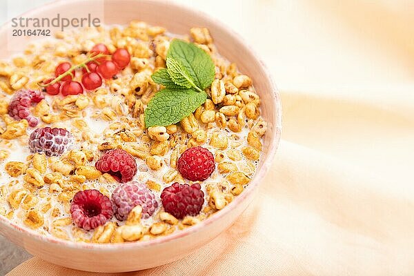 Weizenflockenbrei mit Milch  Himbeeren und Johannisbeeren in einer Keramikschale auf grauem Betonhintergrund und orangefarbenem Leinentextil. Seitenansicht  Nahaufnahme  selektiver Fokus