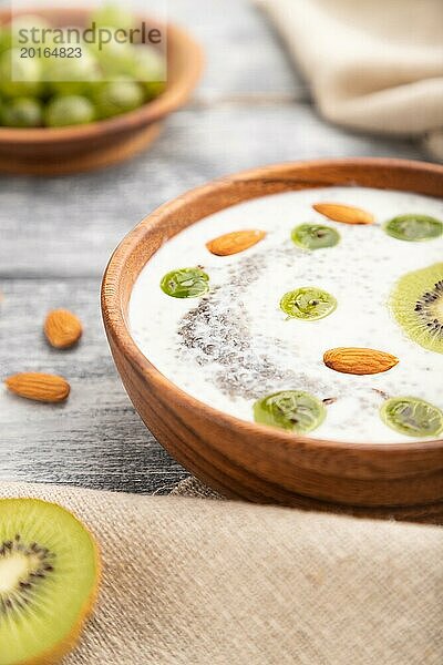 Joghurt mit Kiwi  Stachelbeere  Chia und Mandeln in einer Holzschale auf grauem Holzhintergrund und Leinenstoff. Seitenansicht  Nahaufnahme  selektiver Fokus