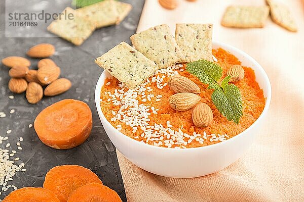 Karottencremesuppe mit Sesam  Mandeln und Knabbereien in weißer Schale auf schwarzem Betonhintergrund mit orangefarbenem Textil. Seitenansicht  Nahaufnahme  selektiver Fokus