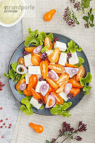 Vegetarischer Salat mit frischen Traubentomaten  Fetakäse  Kopfsalat und Zwiebeln auf blauem Keramikteller auf grauem Betonhintergrund und Leinentextil. Draufsicht  Flachlegung  Nahaufnahme