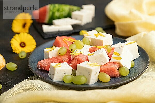 Vegetarischer Salat mit Wassermelone  Fetakäse und Trauben auf blauem Keramikteller auf schwarzem Betonhintergrund und gelbem Leinentuch. Seitenansicht  Nahaufnahme  selektiver Fokus