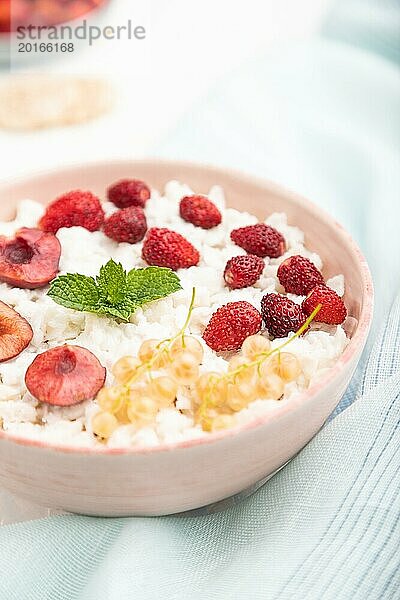 Reisflockenbrei mit Milch und Erdbeeren in Keramikschüssel auf weißem Betonhintergrund und blauem Leinentextil. Seitenansicht  Nahaufnahme  selektiver Fokus