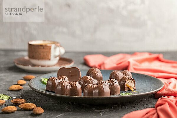 Schokoladenkaramellbonbons mit Mandeln und einer Tasse Kaffee auf einem schwarzen Betonhintergrund und rotem Textil. Seitenansicht  Kopierraum