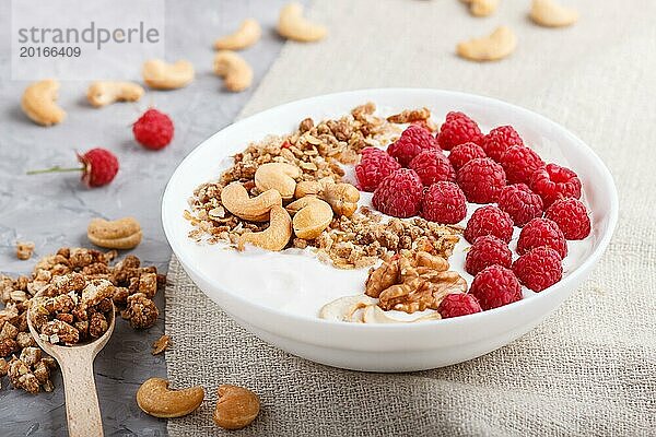 Joghurt mit Himbeere  Müsli  Cashew und Walnuss in weißem Teller mit Holzlöffel auf grauem Betonhintergrund und Leinenstoff. Seitenansicht  Nahaufnahme  selektiver Fokus