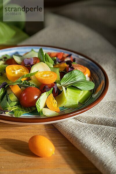 Vegetarischer Salat aus Pac Choi Kohl  Kiwi  Tomaten  Kumquat  mikrogrünen Sprossen auf einem hölzernen Hintergrund und Leinenstoff. Hartes Licht  Kontrast. Seitenansicht  Nahaufnahme  selektiver Fokus