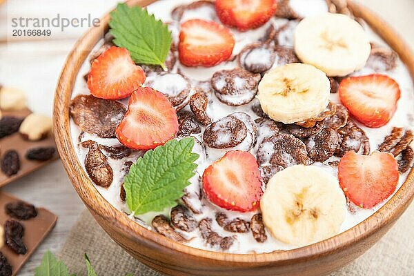 Schokoladen Cornflakes mit Milch und Erdbeeren in einer Holzschale auf weißem Holzhintergrund und Leinenstoff. Seitenansicht  Nahaufnahme  selektiver Fokus