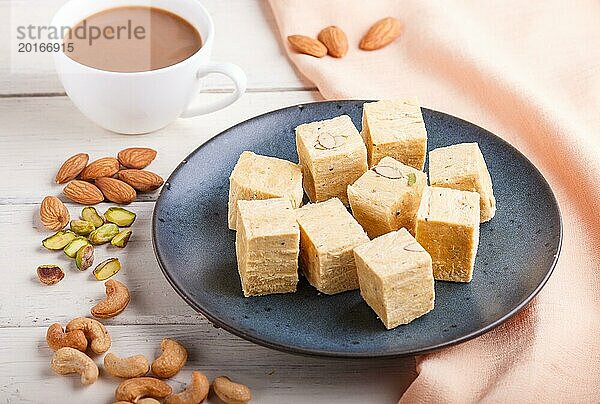 Traditionelle indische Süßigkeit Soan Papdi in einem blauen Keramikteller mit Mandel  Pistache  Cashew und einer Tasse Kaffee auf einem weißen Holzhintergrund mit orangefarbenem Stoff. Seitenansicht  Nahaufnahme  selektiver Fokus