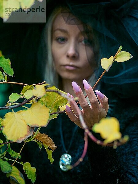 Junge kaukasische blonde dünne blasse Frau in einer nachdenklichen Stimmung mit lebhaften Herbstblättern im Vordergrund  mit Weichzeichner  mit ihrer Hand mit langen manikürten Nägeln  um einen Zweig einer Pflanze zu halten