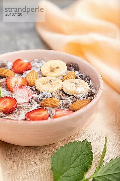 Schokoladen Cornflakes mit Milch  Erdbeeren und Mandeln in einer Keramikschale auf grauem Betonhintergrund und orangefarbenem Leinenstoff. Seitenansicht  Nahaufnahme  selektiver Fokus