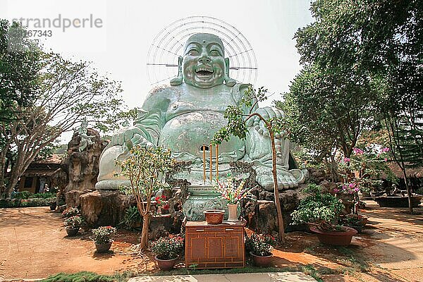 Große lachende Buddha Statue mit Heiligenschein im Freien  umgeben von Bäumen und einem Altar mit Blumen  Linh An Pagode. Dalat  Vietnam  Asien