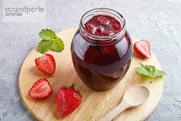 Erdbeermarmelade in einem Glas mit Beeren und Blättern auf grauem Betonhintergrund. Hausgemacht  Nahaufnahme  Seitenansicht  selektiver Fokus