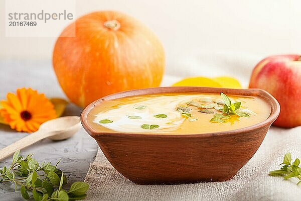 Traditionelle Kürbiscremesuppe mit Kernen in einer Tonschale auf grauem Betonhintergrund mit Leinentextil. Seitenansicht  Nahaufnahme  selektiver Fokus