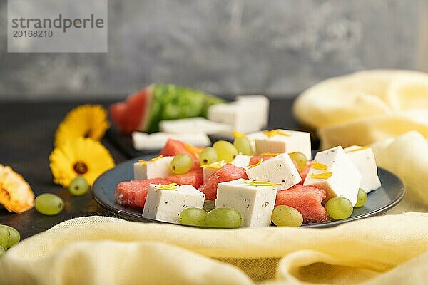 Vegetarischer Salat mit Wassermelone  Fetakäse und Trauben auf blauem Keramikteller auf schwarzem Betonhintergrund und gelbem Leinentuch. Seitenansicht  Nahaufnahme  selektiver Fokus