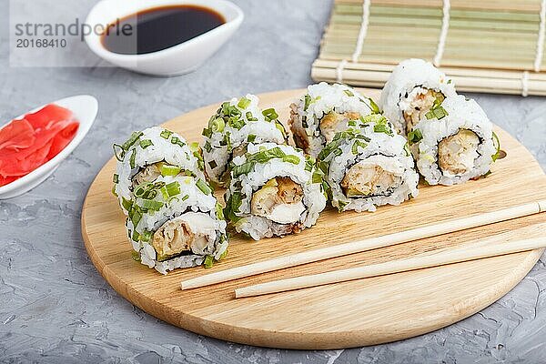 Japanische Maki Sushi Rollen mit grünen Zwiebeln  Essstäbchen  Sojasauce und mariniertem Ingwer auf einem Holzbrett vor einem grauen Betonhintergrund. Seitenansicht  Nahaufnahme  selektiver Fokus