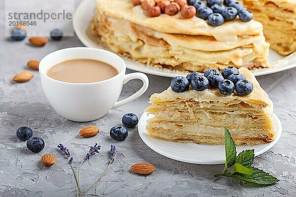 Hausgemachte geschichteten Napoleon Kuchen mit Milch Creme. Dekoriert mit Heidelbeere  Mandeln  Walnüsse  Haselnüsse  Minze auf einem grauen Beton Hintergrund und Tasse Kaffee. Seitenansicht  Nahaufnahme  selektiver Fokus