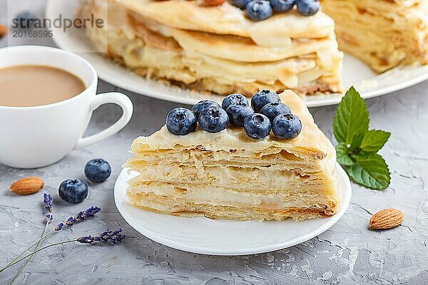 Hausgemachte geschichteten Napoleon Kuchen mit Milch Creme. Dekoriert mit Heidelbeere  Mandeln  Walnüsse  Haselnüsse  Minze auf einem grauen Beton Hintergrund und Tasse Kaffee. Seitenansicht  Nahaufnahme  selektiver Fokus