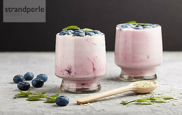 Joghurt mit Blaubeeren und Sesam im Glas und Holzlöffel auf grauem und schwarzem Hintergrund. Seitenansicht  Nahaufnahme