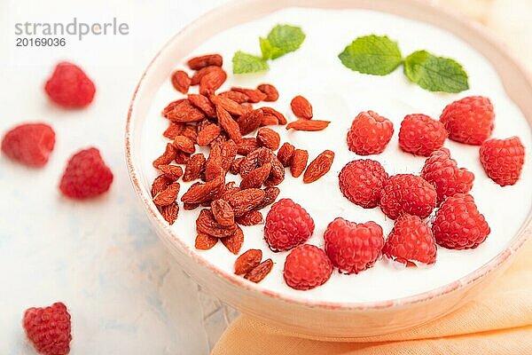 Joghurt mit Himbeeren und Goji Beeren in einer Keramikschale auf weißem Betonhintergrund und orangefarbenem Leinentextil. Seitenansicht  Nahaufnahme  selektiver Fokus