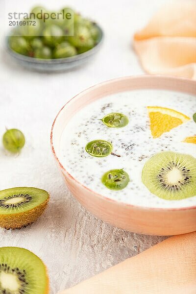 Joghurt mit Kiwi  Stachelbeere und Chia in einer Keramikschale auf grauem Betonhintergrund und orangefarbenem Leinentextil. Seitenansicht  Nahaufnahme  selektiver Fokus