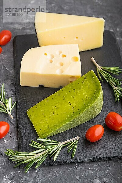 Grüner Basilikumkäse und verschiedene Käsesorten mit Rosmarin und Tomaten auf einer schwarzen Schiefertafel auf einem schwarzen Betonhintergrund. Seitenansicht  Nahaufnahme  selektiver Fokus