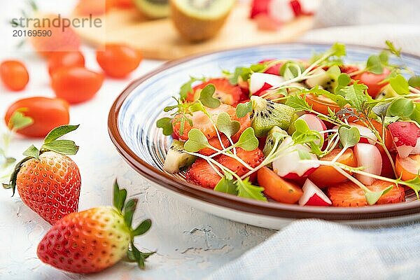 Vegetarischer Obst und Gemüsesalat aus Erdbeeren  Kiwi  Tomaten  Mikrogrünsprossen auf weißem Betonhintergrund und Leinentextil. Seitenansicht  Nahaufnahme  selektiver Fokus