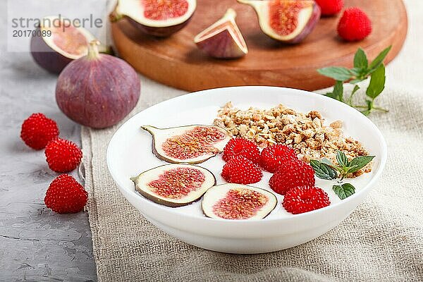 Joghurt mit Himbeeren  Müsli und Feigen in einem weißen Teller auf einem grauen Betonhintergrund und Leinentextil. Seitenansicht  Nahaufnahme  selektiver Fokus