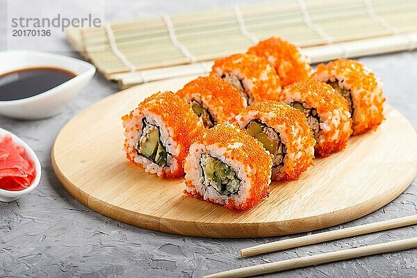 Japanische Maki Sushi Rollen mit fliegendem Fischrogen  Essstäbchen  Sojasauce und mariniertem Ingwer auf einem Holzbrett vor einem grauen Betonhintergrund. Seitenansicht  Nahaufnahme  selektiver Fokus