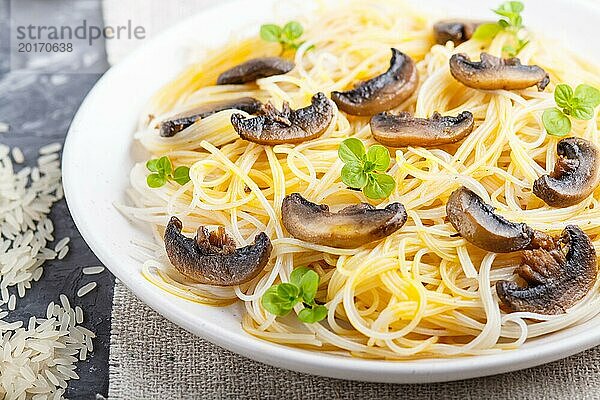 Reisnudeln mit Champignons  Eiersauce und Oregano auf weißem Keramikteller vor schwarzem Betonhintergrund. Seitenansicht  selektiver Fokus  Nahaufnahme