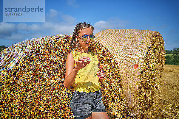 Mädchen trägt Sonnenbrille und stützt sich auf einen Strohballen im Feld