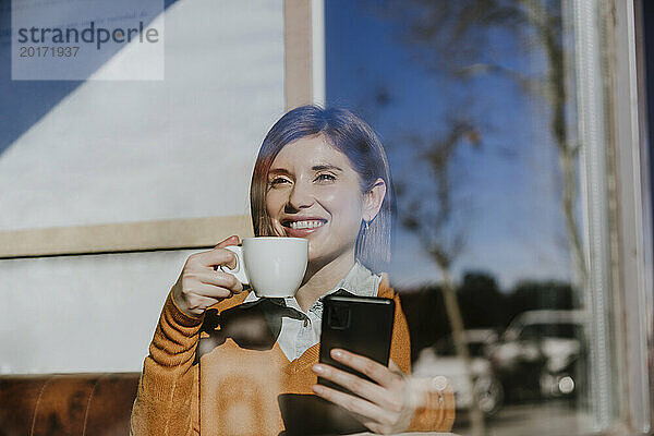 Glückliche Frau sitzt mit Smartphone und Kaffeetasse im Café