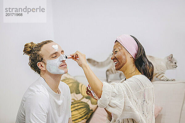 Glückliche Frau trägt zu Hause eine Hautpflegemaske auf das Gesicht ihres Freundes auf