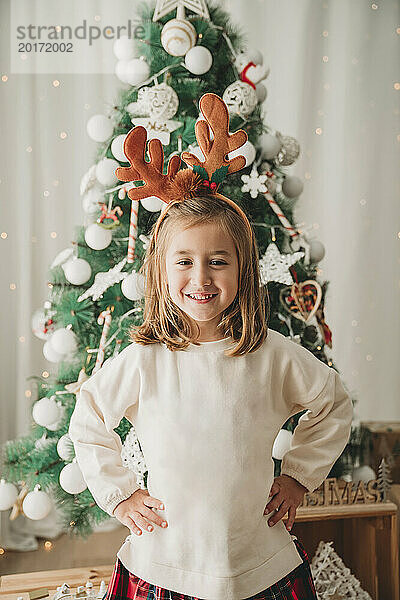 Glückliches Mädchen  das mit den Händen auf der Hüfte vor dem Weihnachtsbaum steht
