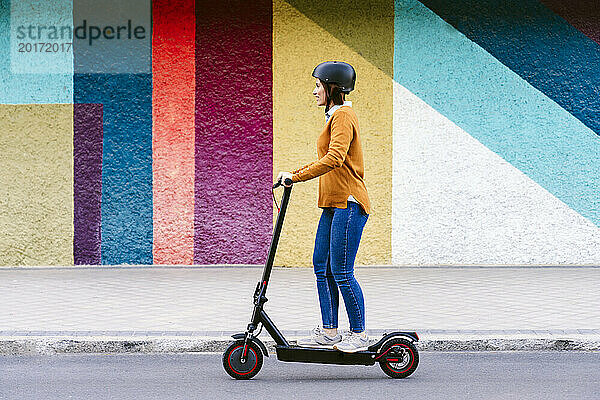 Frau trägt Helm und fährt Elektroroller vor bunter Wand