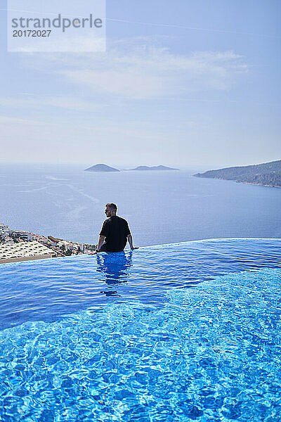 Carefree man sitting at the edge of blue swimming pool in villa