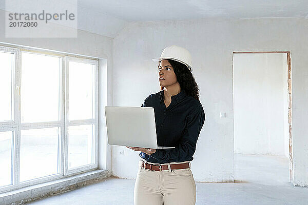 Architekt trägt Schutzhelm und steht mit Laptop vor Ort