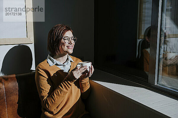 Smiling woman holding coffee cup and enjoying sunlight in cafe