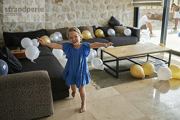 Fröhliches Mädchen steht mit ausgestreckten Armen und Geburtstagsballons im Wohnzimmer der Villa