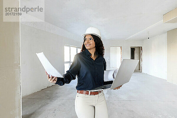 Lächelnder junger Architekt hält Laptop in der Hand und untersucht die Baustelle mit Bauplänen