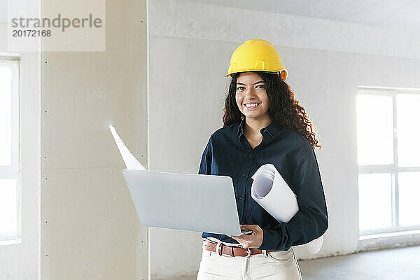 Lächelnder junger Architekt hält Laptop und Baupläne vor Ort