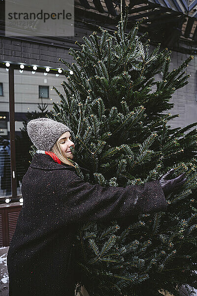 Lächelnde Frau mit Strickmütze und Weihnachtsbaum umarmend