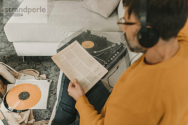 Man listening to music through headphones reading paper sitting by record player on sofa at home