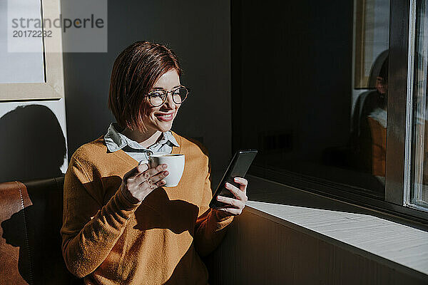 Lächelnde Frau hält Kaffeetasse in der Hand und benutzt Smartphone im Café