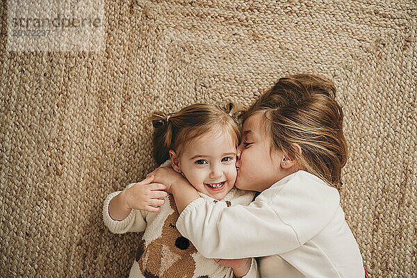 Mädchen küsst lächelnde Schwester  die zu Hause auf dem Teppich liegt