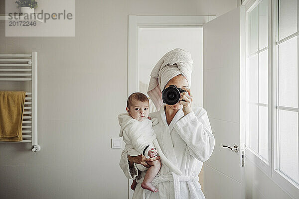 Mutter trägt Baby und fotografiert mit der Kamera im Badezimmer zu Hause