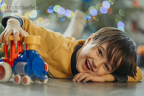 Fröhlicher Junge liegt auf dem Boden und spielt zu Hause mit Zugspielzeug
