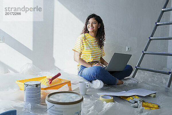 Lächelnde Frau sitzt mit Laptop auf dem Boden in einem renovierten Zimmer